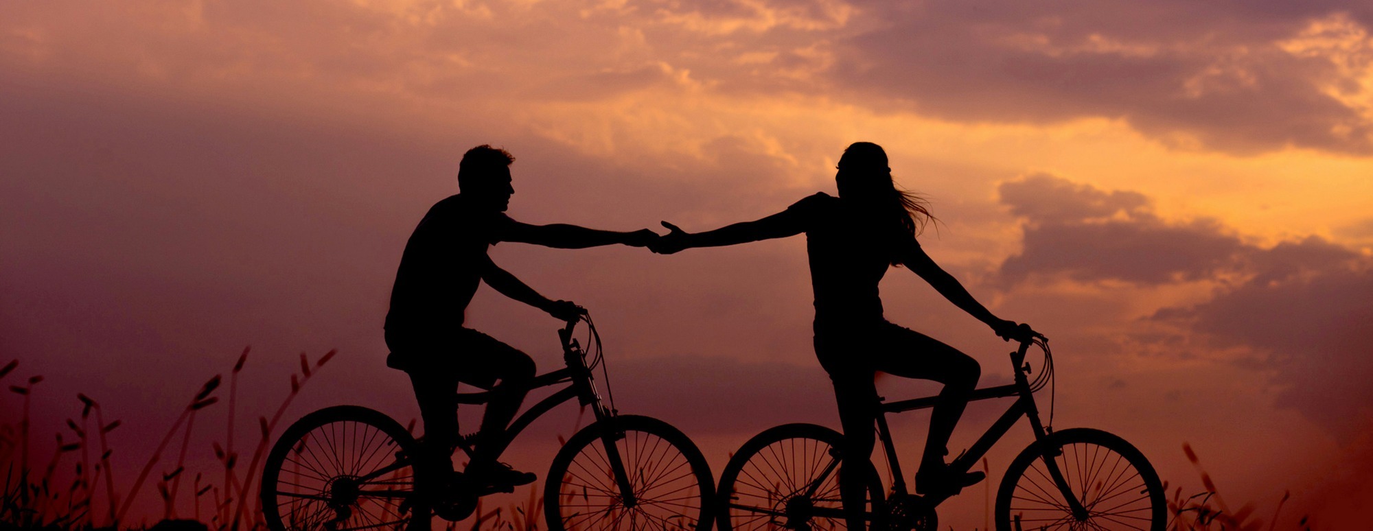 Doi oameni pe biciclete la apus, atingându-și mâinile — simbol al conexiunii și atașamentului